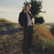 model stands on a dirt path lined by red flowers and overlooking a view wearing a lavender button up, a sweater, and trousers